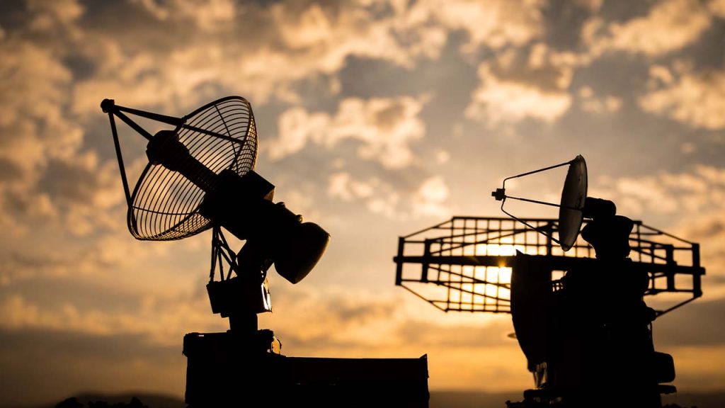 Silhouettes of two radar or satellite dishes against a dramatic, cloudy sky at sunset. The dish on the left has a mesh design, and the one on the right has a rectangular grid structure.