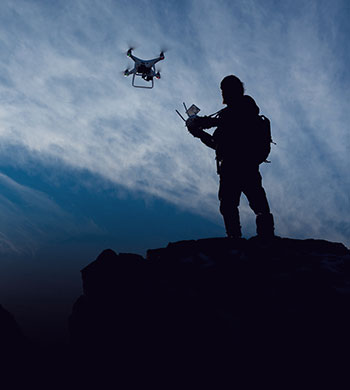 Silhouette of a person controlling a drone on a rocky cliff. The sky is a gradient of blue, creating a dramatic and adventurous atmosphere. The person is holding a remote control and carrying a backpack.