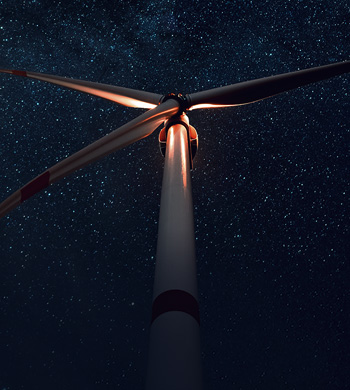 View from the base of a large wind turbine at night, illuminated by lights. The turbine's blades are prominent against a clear, starry sky, creating a striking contrast between technology and nature.