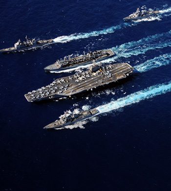 An aerial view of an aircraft carrier surrounded by three smaller naval ships, all moving through a deep blue ocean, leaving trails of white wake behind. The aircraft carrier has numerous aircraft on its deck.