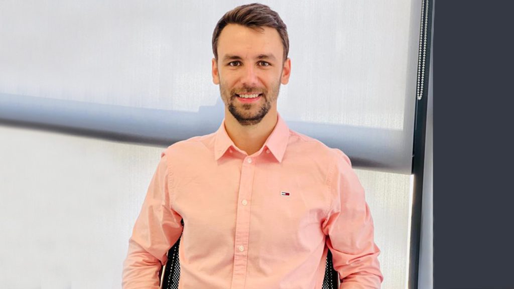 A man with short brown hair and a beard, wearing a light pink button-up shirt, sits on a black chair in front of a window with blinds, smiling at the camera.