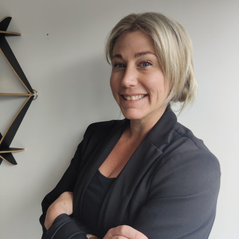 A woman with light blonde hair in a loose bun, wearing a black blazer and top, stands smiling with arms crossed in front of a plain light-colored wall with part of a geometric shelf visible on the left.