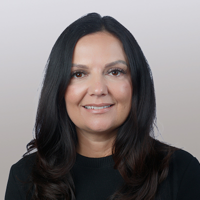 A woman with long dark hair and brown eyes, wearing a black top, smiles at the camera against a light, gradient background.