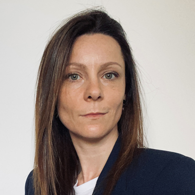 A woman with straight brown hair and light skin wearing a dark blazer and white top, facing the camera with a neutral expression against a plain light background.