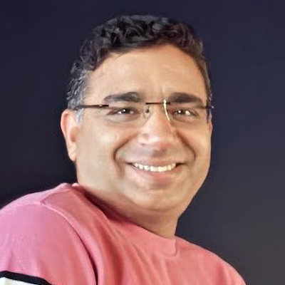 A man with short, curly dark hair and glasses smiles at the camera. He is wearing a pink shirt and is set against a dark background.