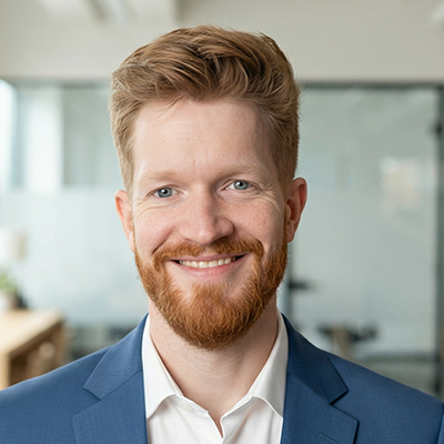 A man with light skin, short red hair, and a full red beard is smiling. He is wearing a blue suit jacket and a white shirt, standing in a bright office with glass walls in the background.