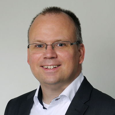 A man with short brown hair and glasses is smiling at the camera. He is wearing a white shirt with a dark suit jacket. The background is plain and light-colored.